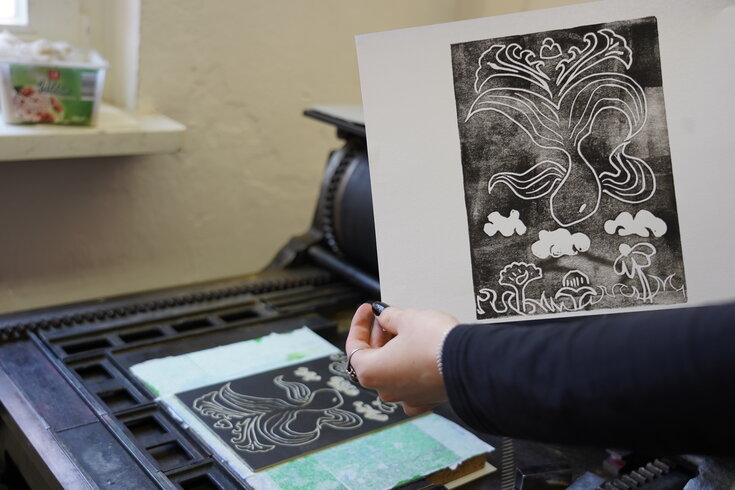 Bedrucktes Bild wird leicht hochgehalten zum Vorzeigen. Siebdruckmaschine im Hintergrund.