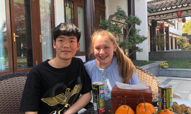 Eine deutsche Schülerin und ein chinesischer Schüler sitzen im Freien an einem Tisch vor einer Schale mit Orangen