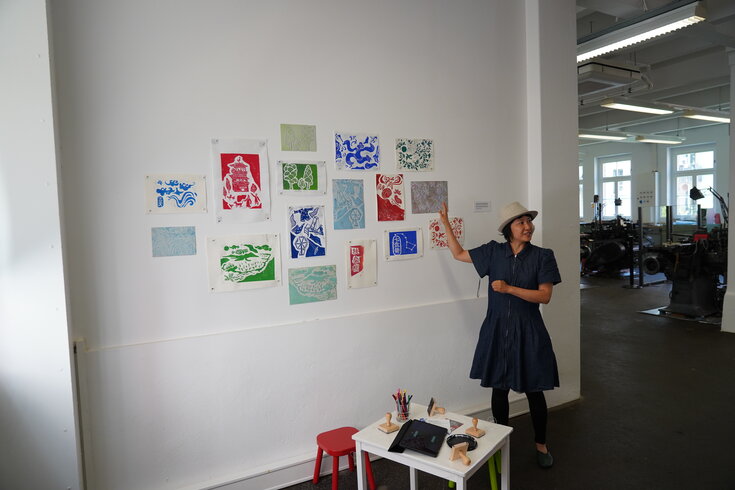 Ergebnisse des Druckworkshops hängen an der Wand. Leiterin steht davor und spricht. 