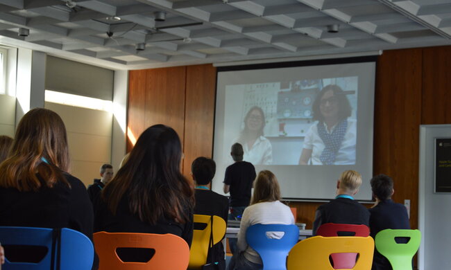 Ein Kurs mit Schüler:innen blickt auf eine Leinwand mit einem Videocall